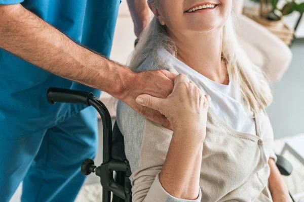 Plan recadré de femme âgée souriante en fauteuil roulant tenant la main de l'aidant — Photo de stock