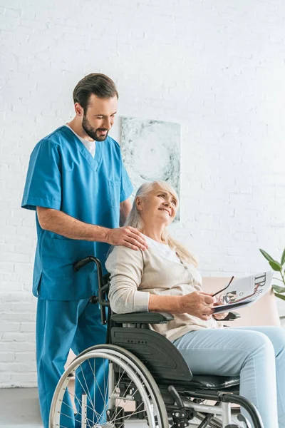 Sorridente caregiver guardando felice anziana donna leggendo giornale in sedia a rotelle — Foto stock