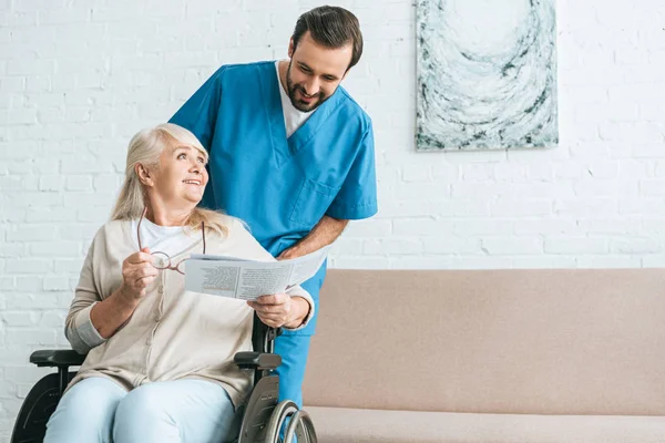 Sorridente giovane infermiera maschile guardando felice donna anziana leggere giornale in sedia a rotelle — Foto stock