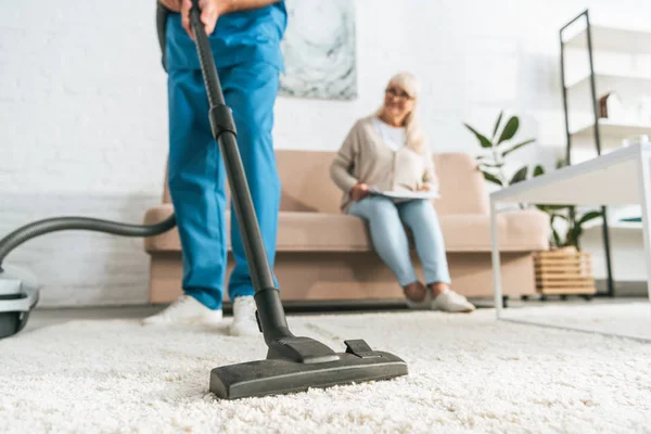 Cropped shot of man using vacuum cleaner while senior woman sitting on sofa — Stock Photo