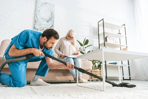 Jeune homme utilisant aspirateur tandis que la femme âgée assis sur le canapé — Photo de stock