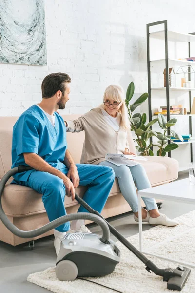 Sorridente donna anziana seduta sul divano e guardando caregiver con aspirapolvere — Foto stock