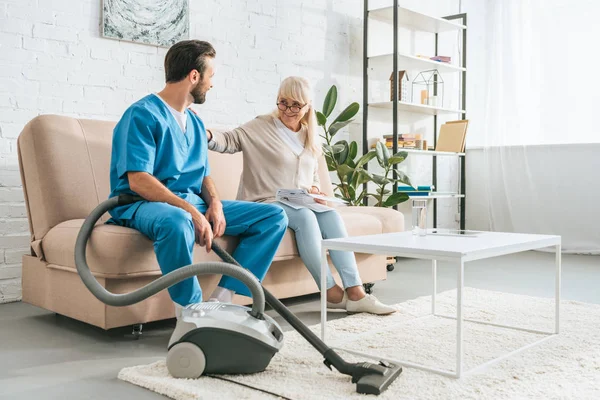 Femme âgée souriante assise sur le canapé et regardant travailleur social avec aspirateur — Photo de stock