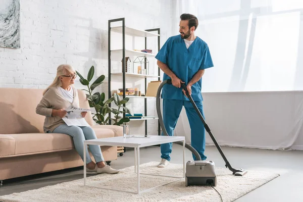 Smiling social worker cleaning carpet with vacuum cleaner and looking at senior woman reading newspaper — Stock Photo