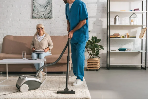 Schnappschuss von Sozialarbeiterin, die Teppich mit Staubsauger reinigt, während Seniorin Zeitung liest — Stockfoto