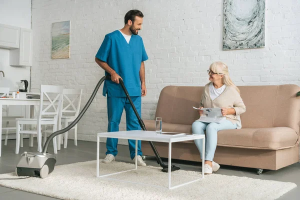 Smiling social worker using vacuum cleaner and looking at senior woman reading newspaper on couch — Stock Photo