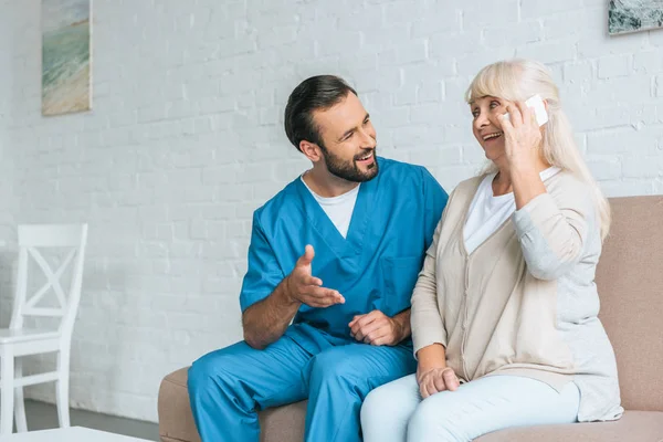 Lächelnder Krankenpfleger schaut glückliche Seniorin an, die per Smartphone spricht — Stockfoto