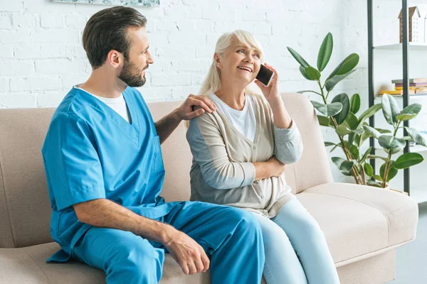 Giovane badante guardando sorridente donna anziana che parla da smartphone — Foto stock