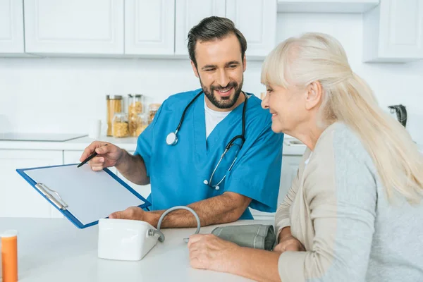 Enfermera masculina sonriente mostrando portapapeles a mujer mayor feliz después de medir la presión arterial - foto de stock