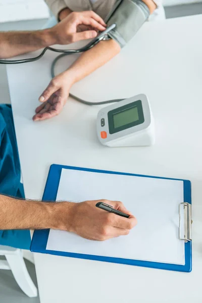 Recortado tiro de enfermera masculina escribir en portapapeles mientras que la medición de la presión arterial a la mujer mayor - foto de stock