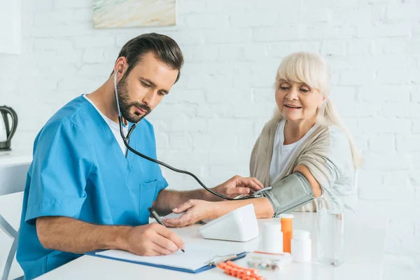 Krankenpfleger in Stethoskop schreibt auf Klemmbrett, während er den Blutdruck einer Seniorin misst — Stockfoto