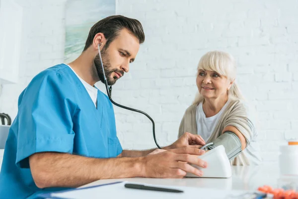 Visión de ángulo bajo de la enfermera masculina que mide la presión arterial a la mujer mayor sonriente - foto de stock