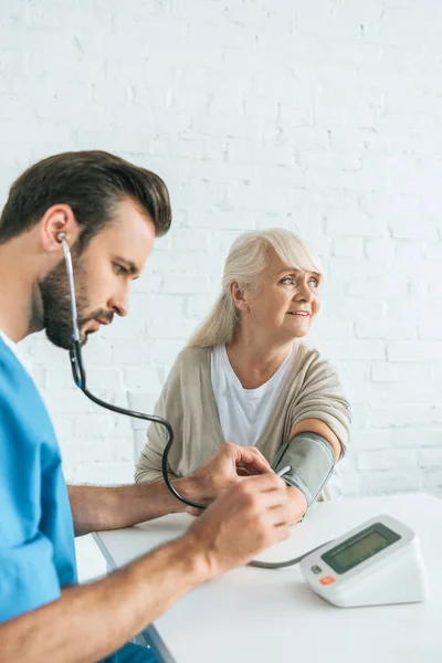 Male nurse in stethoscope measuring blood pressure to smiling senior woman — Stock Photo