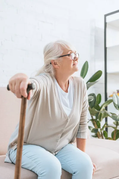 Seniorin mit Brille hält Gehstock in der Hand und schaut weg, während sie auf Sofa sitzt — Stockfoto