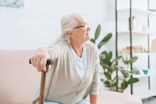 Senior woman in eyeglasses holding walking stick and looking away while sitting on couch — Stock Photo