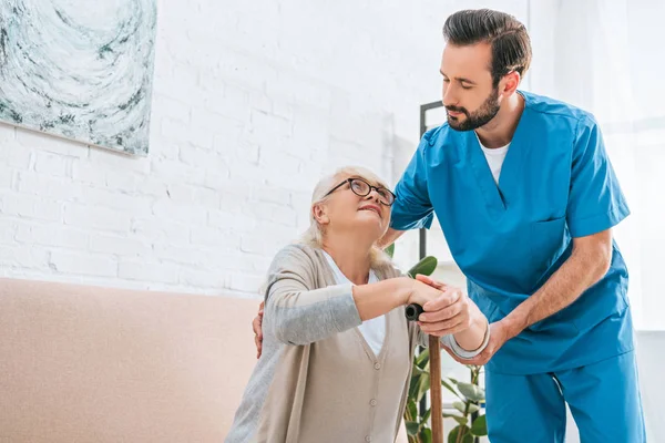 Young male social worker supporting senior woman in eyeglasses with walking stick — Stock Photo