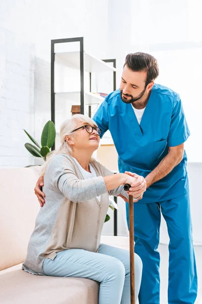 Giovane infermiera di sesso maschile che sostiene la donna anziana negli occhiali con bastone da passeggio — Foto stock