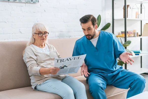Chocado enfermeira masculina olhando para a mulher sênior em óculos lendo jornal de viagem — Fotografia de Stock