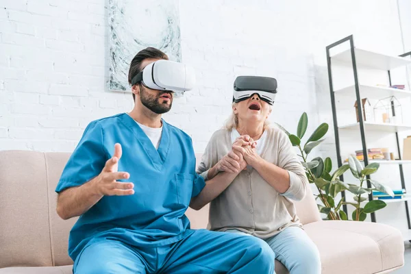 Donna anziana e giovane infermiera maschile utilizzando cuffie realtà virtuale e tenendosi per mano — Stock Photo