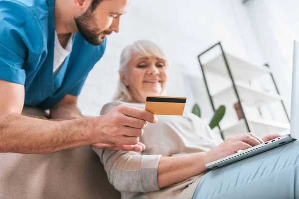 Assistente social segurando cartão de crédito e olhando para a mulher idosa usando laptop — Fotografia de Stock