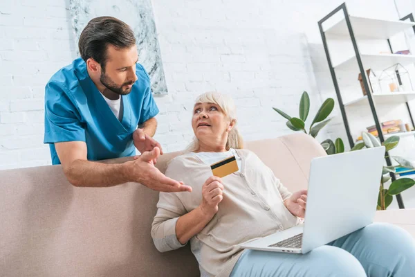 Infirmière masculine regardant une femme âgée tenant une carte de crédit et utilisant un ordinateur portable — Photo de stock