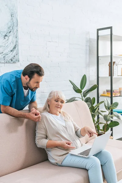 Чоловік медсестра спирається на диван і дивиться на старшу жінку, використовуючи ноутбук — стокове фото