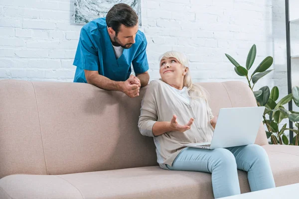 Joven enfermera masculina apoyada en el sofá y mirando a la mujer mayor utilizando el ordenador portátil - foto de stock