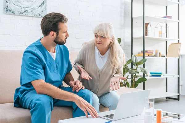 Senior woman and young caregiver looking at each other while using laptop together — Stock Photo