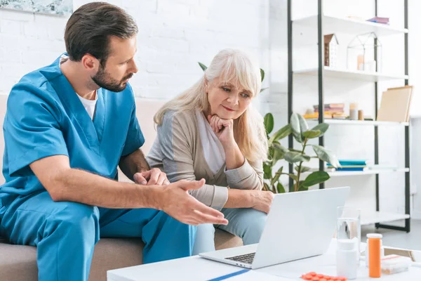 Smiling senior woman looking at laptop while sitting on couch with social worker — Stock Photo