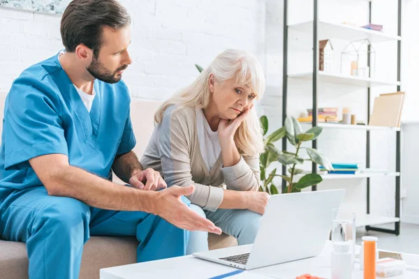 Molesto mujer mayor mirando a la computadora portátil mientras está sentado en el sofá con el trabajador social - foto de stock