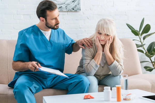 Male nurse holding clipboard and supporting upset senior woman looking at containers with pills — Stock Photo