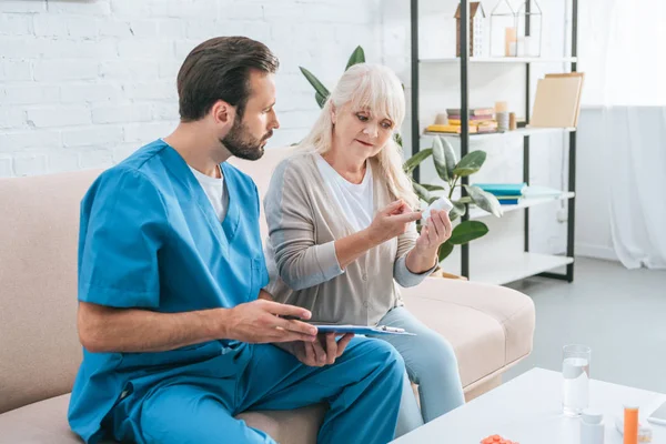 Male nurse with clipboard looking at senior woman holding pills — Stock Photo