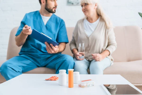 Vista close-up de recipientes com pílulas e tablet digital na mesa, enfermeiro e mulher idosa sentados no sofá atrás — Fotografia de Stock