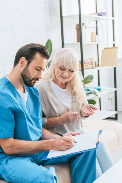 Seniorin nimmt Tabletten und betrachtet Krankenschwester, die auf Klemmbrett schreibt — Stockfoto