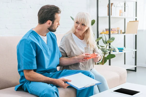 Junge männliche Krankenschwester hält Klemmbrett und gibt der Seniorin ein Glas Wasser mit Pillen — Stockfoto