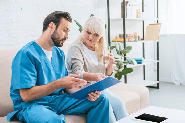 Junge männliche Krankenschwester zeigt Klemmbrett der Seniorin mit einem Glas Wasser — Stockfoto