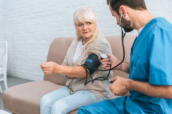 Social worker measuring blood pressure to senior woman sitting on couch — Stock Photo