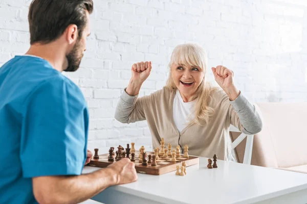 Triumphierende Seniorin feiert Sieg und schaut junge Sozialarbeiterin nach Schachspiel an — Stockfoto