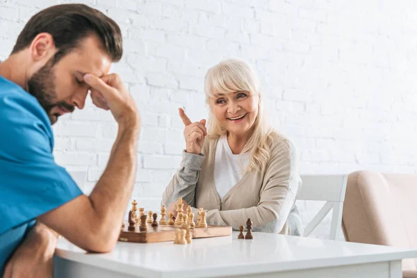 Sorrindo mulher sênior apontando com o dedo e olhando para o homem chateado após jogo de xadrez — Fotografia de Stock