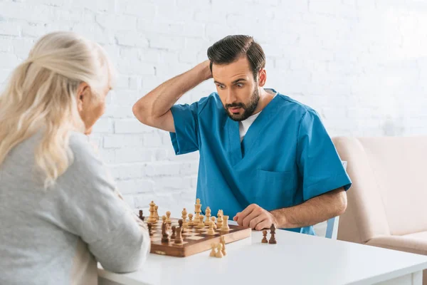 Confus soignant masculin jouant aux échecs avec une femme âgée — Photo de stock