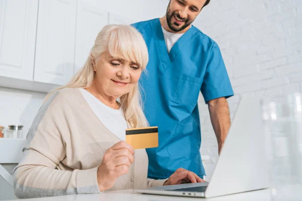 Assistente sociale sorridente guardando donna anziana in possesso di carta di credito e utilizzando il computer portatile — Foto stock