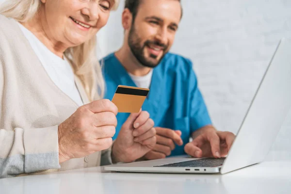 Plan recadré d'une femme âgée souriante tenant une carte de crédit et utilisant un ordinateur portable avec un jeune travailleur social — Photo de stock