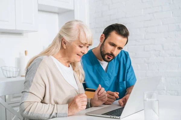 Smiling senior woman holding credit card and using laptop with young caregiver — Stock Photo