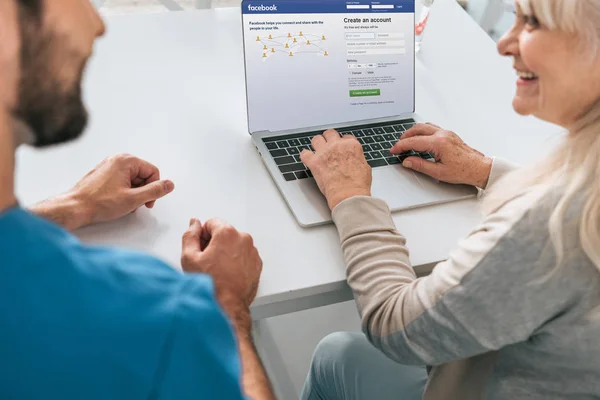 Cropped shot of young man and smiling senior woman using laptop with facebook website on screen — Stock Photo