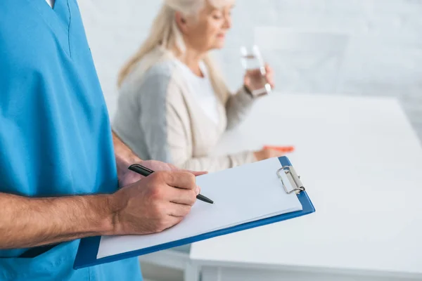 Vista parcial de cerca de la escritura del cuidador en el portapapeles mientras la mujer mayor toma medicamentos - foto de stock