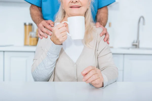 Recortado disparo de trabajador social de pie cerca de la mujer mayor beber té en la cocina - foto de stock