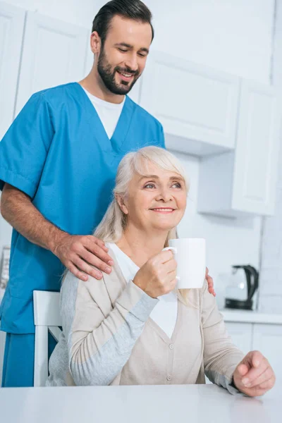 Sorridente badante maschile guardando la donna anziana bere il tè in cucina — Foto stock