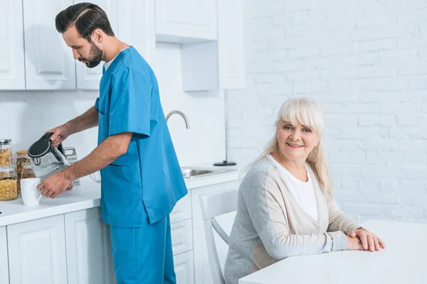 Glückliche Seniorin sitzt lächelnd in die Kamera, während Betreuerin Heißgetränk in Küche einschenkt — Stockfoto