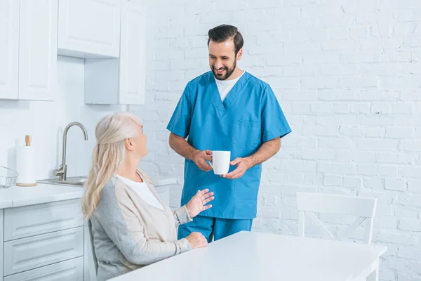 Cuidador sonriente sosteniendo la taza y mirando a la mujer mayor sentada en la cocina - foto de stock