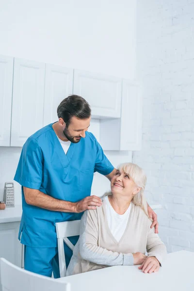 Feliz anciano mujer y joven cuidador sonriendo entre sí - foto de stock